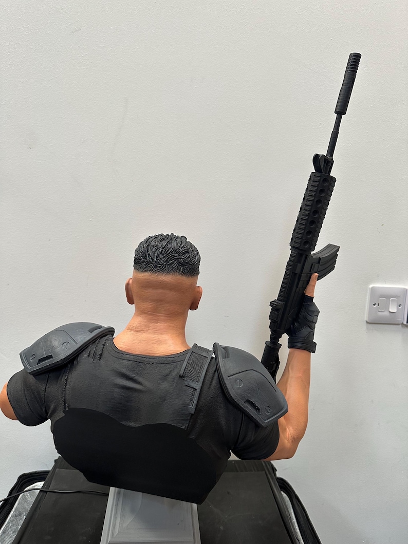 May include: A detailed bust of a figure in a black tactical shirt and shoulder pads, holding a black rifle. The figure's hair is neatly styled, and the overall design suggests a military or action theme.