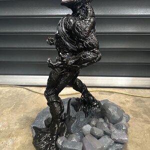 May include: A black Venom statue with a white stripe on the chest and a red tongue. The statue is standing on a gray base with gray rocks. The figure has a detailed, textured appearance.