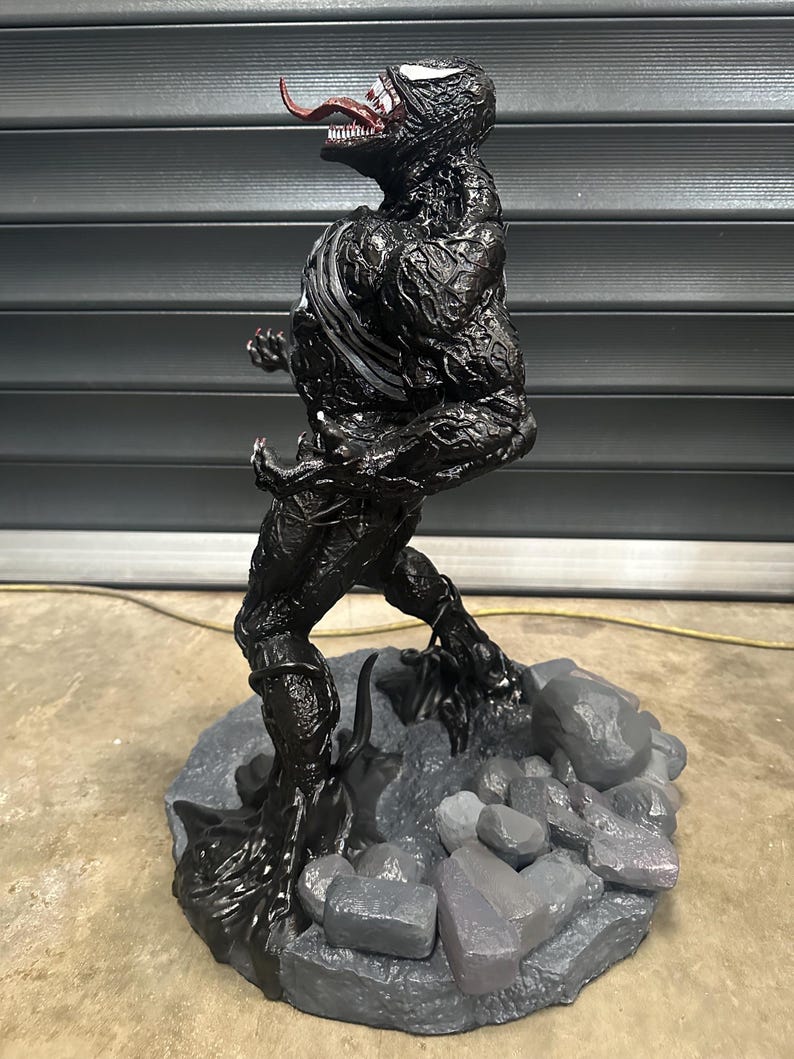 May include: A black Venom statue with a white stripe on the chest and a red tongue. The statue is standing on a gray base with gray rocks. The figure has a detailed, textured appearance.