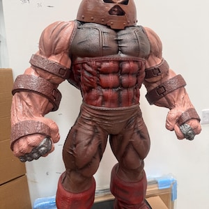 May include: A detailed figurine of the Juggernaut, a Marvel Comics character. The figure is predominantly brown and red, with a helmet, armored chest, and muscular physique. The figure is standing upright, showcasing its powerful build.