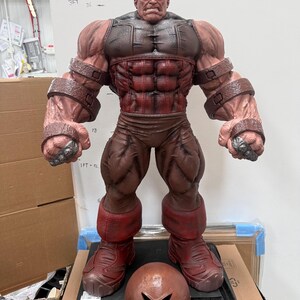 May include: A large, detailed figurine of a muscular character with red and brown tones. The figure has a stern expression and is wearing a helmet. The figurine is standing, with a helmet placed in front of it.
