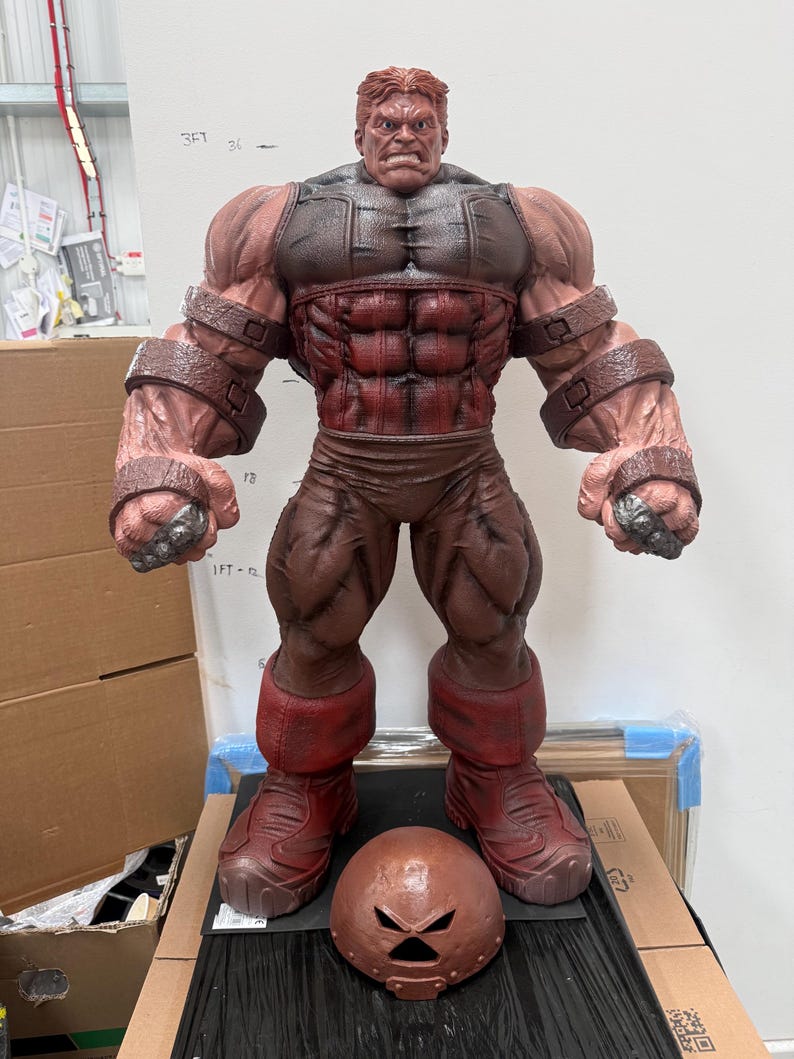 May include: A large, detailed figurine of a muscular character with red and brown tones. The figure has a stern expression and is wearing a helmet. The figurine is standing, with a helmet placed in front of it.