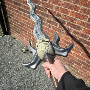 May include: A fantasy sword with a curved, gray blade and a detailed hilt. The hilt features a skull-like design and is held by a brown handle. The sword is held against a brick wall background.