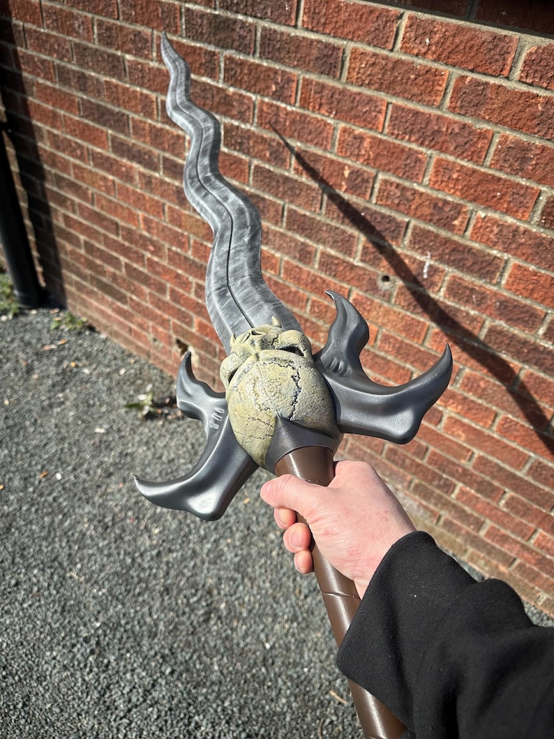 May include: A fantasy sword with a curved, gray blade and a detailed hilt. The hilt features a skull-like design and is held by a brown handle. The sword is held against a brick wall background.