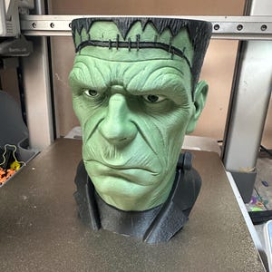 Puede incluir: Busto verde y negro del monstruo de Frankenstein. El busto tiene una cara verde con una expresi&oacute;n seria, cabello negro y una chaqueta negra. Es un art&iacute;culo decorativo.