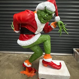 May include: A large, green Grinch figure in a Santa Claus outfit, poised on a chimney. The Grinch wears a red Santa suit with white trim, a matching hat, and red shoes. The figure is a festive Christmas decoration.