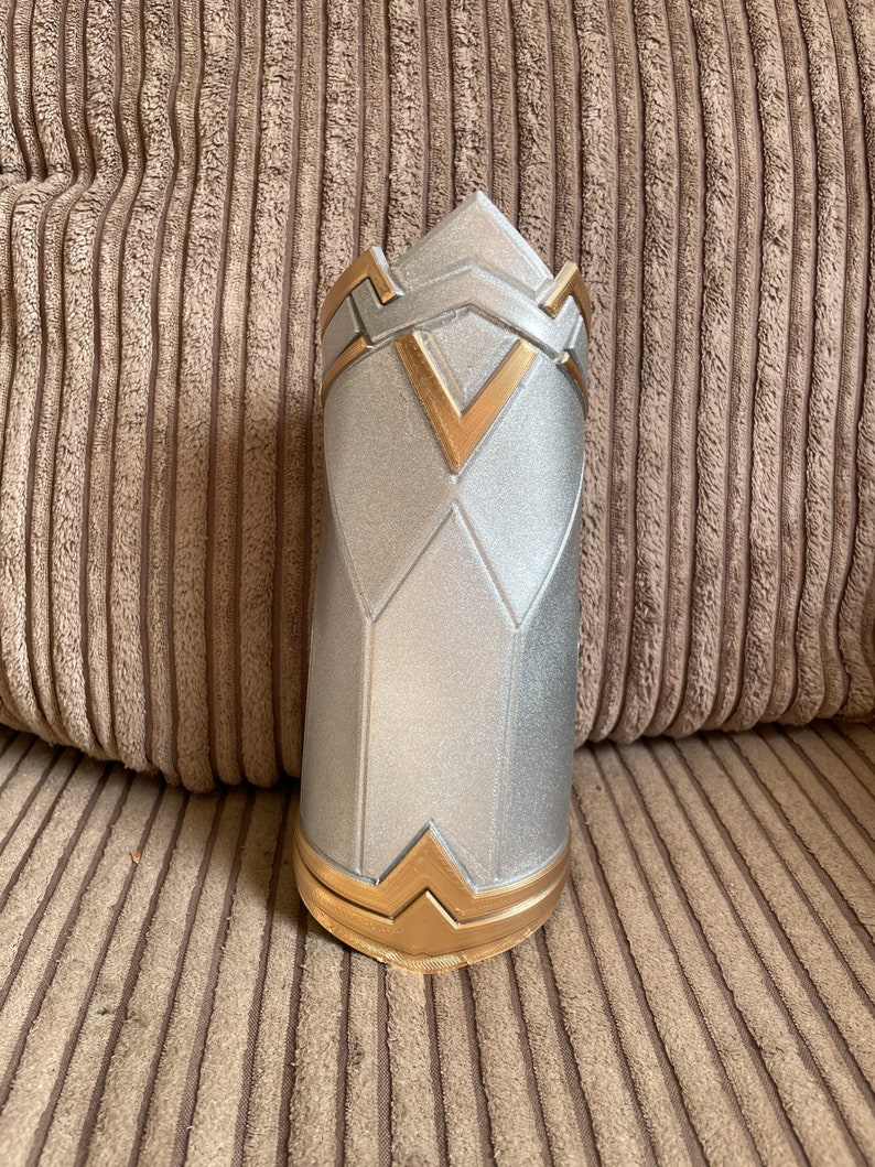 May include: A silver and gold decorative arm cuff with a geometric design. The cuff is made of a hard material and has a smooth finish.