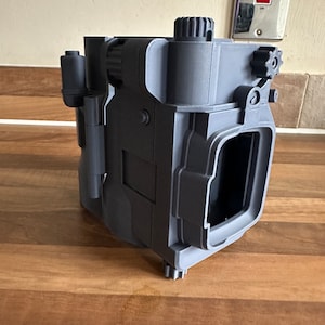 May include: A 3D printed grey plastic model of a camera with a gear on the top and side. The camera has a rectangular shape with a square opening on the front.