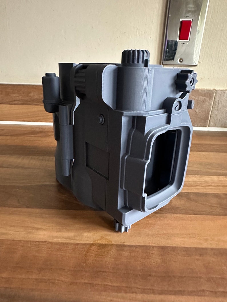 May include: A 3D printed grey plastic model of a camera with a gear on the top and side. The camera has a rectangular shape with a square opening on the front.
