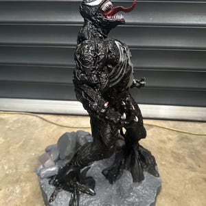 May include: A black Venom action figure with a red tongue and white teeth. The figure has a detailed texture and stands on a gray rocky base. The figure's chest features white accents.