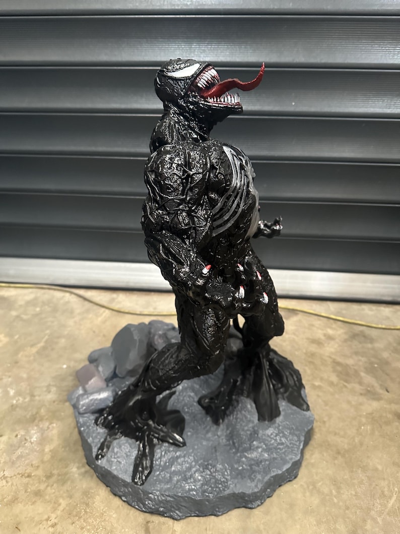 May include: A black Venom action figure with a red tongue and white teeth. The figure has a detailed texture and stands on a gray rocky base. The figure's chest features white accents.