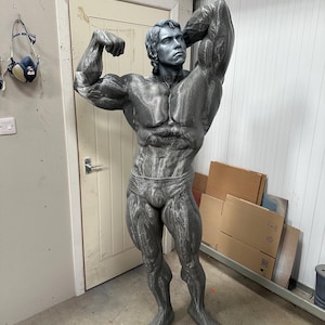 May include: A gray sculpture of a bodybuilder in a classic pose, flexing his biceps. The statue is detailed with realistic muscle definition and is wearing briefs. The sculpture is standing in a workshop setting.