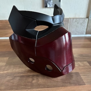 May include: A burgundy and black plastic mask with a geometric design. The mask has two eye holes and a nose hole.