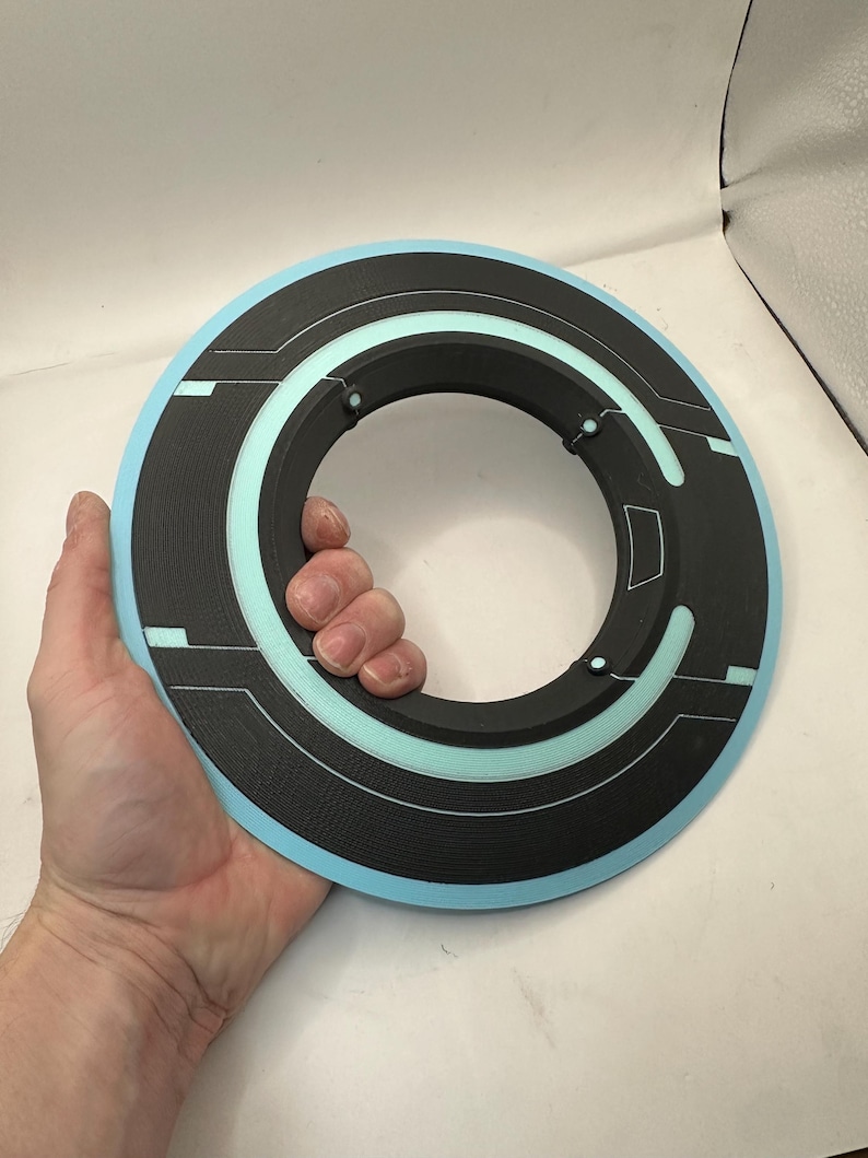 May include: A black and light blue circular prop with a textured surface. The prop has a ring shape with a central opening and a light blue outer edge. The design includes linear and curved details.