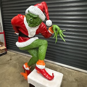 May include: A large, green Grinch statue wearing a red Santa suit and hat with white trim. The Grinch is posed on a white chimney, with orange accents on the boots and gloves. The statue is a festive Christmas decoration.