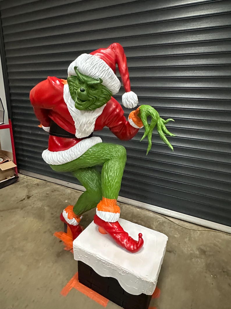 May include: A large, green Grinch statue wearing a red Santa suit and hat with white trim. The Grinch is posed on a white chimney, with orange accents on the boots and gloves. The statue is a festive Christmas decoration.