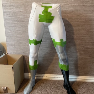 Puede incluir: Una forma de pierna de maniqu&iacute; blanca y gris con detalles de cinta verde. Una pierna es negra. La forma est&aacute; sobre una alfombra marr&oacute;n con una caja de cart&oacute;n en el fondo. La forma es probablemente para exhibir ropa.