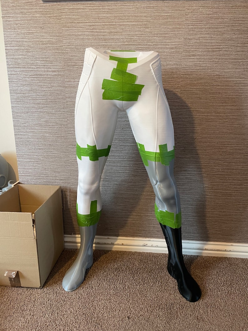 Puede incluir: Una forma de pierna de maniqu&iacute; blanca y gris con detalles de cinta verde. Una pierna es negra. La forma est&aacute; sobre una alfombra marr&oacute;n con una caja de cart&oacute;n en el fondo. La forma es probablemente para exhibir ropa.