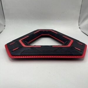 May include: A black and red geometric object with a hexagonal shape. The object has a textured surface with red accents and a honeycomb-like pattern on the bottom edge. The design is symmetrical and appears to be 3D-printed.