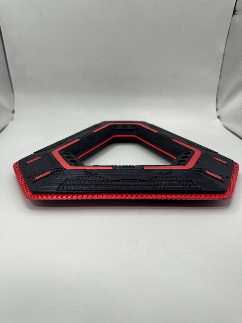 May include: A black and red geometric object with a hexagonal shape. The object has a textured surface with red accents and a honeycomb-like pattern on the bottom edge. The design is symmetrical and appears to be 3D-printed.