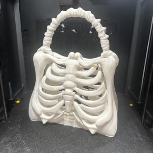 May include: A white, 3D-printed handbag in the shape of a rib cage, with a handle made of vertebrae. The bag is on a dark surface, possibly inside a 3D printer. The design is detailed, showcasing a unique, gothic aesthetic. The bag is a statement piece.