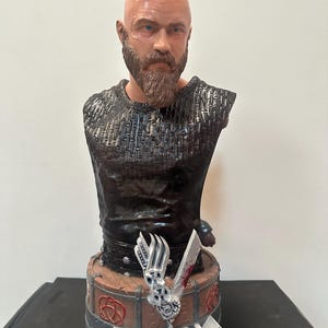May include: A detailed bust of a Viking warrior with a shaved head and a full beard. The bust is dark with intricate armour detailing. The base features the word "VIKING" and a decorative design with a sword.