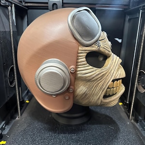 May include: A brown and silver helmet with a detailed face design, reminiscent of a skull, is displayed inside a 3D printer. The helmet has a vintage aviator style with goggles and circular side accents. The face is tan with visible teeth.