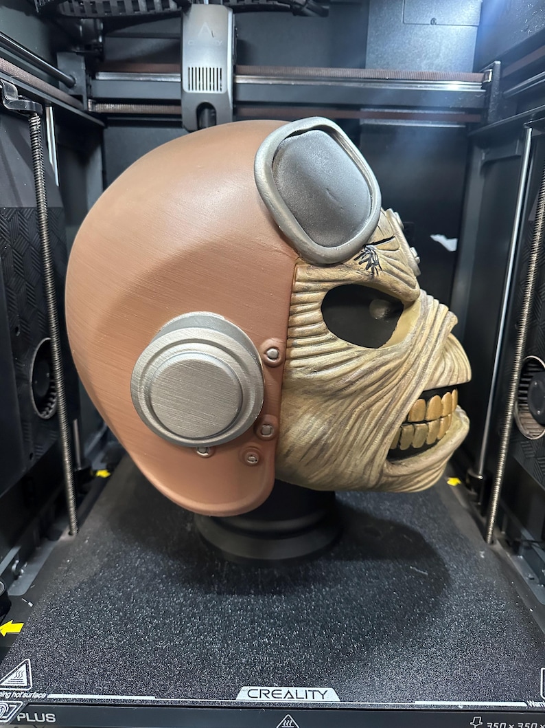 May include: A brown and silver helmet with a detailed face design, reminiscent of a skull, is displayed inside a 3D printer. The helmet has a vintage aviator style with goggles and circular side accents. The face is tan with visible teeth.