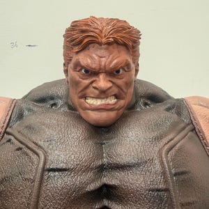 May include: A detailed action figure of a muscular character with a stern expression. The figure has red hair, a brown complexion, and is wearing a brown and tan outfit. The figure's face is contorted in an angry grimace.