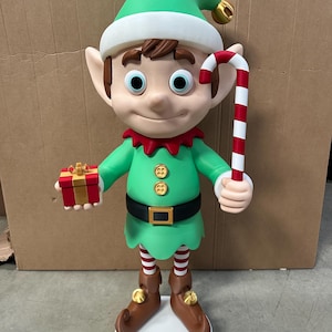 May include: A large, festive elf figurine with a green hat, tunic, and shoes. The elf holds a candy cane and a small gift box. The elf's belt has a gold buckle, and its striped socks add a playful touch. A perfect Christmas decoration.