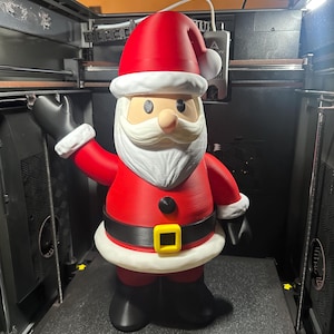 May include: A 3D-printed Santa Claus figure, featuring a red suit and hat with white trim, a white beard, and black gloves and boots. The figure has a yellow belt buckle and is standing in a 3D printer.
