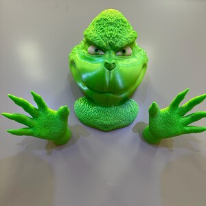 May include: A green Grinch figure with a mischievous expression, with two green hands outstretched. The figure is made of plastic and has a textured surface.