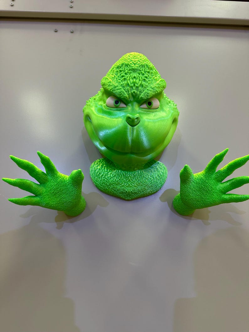 May include: A green Grinch figure with a mischievous expression, with two green hands outstretched. The figure is made of plastic and has a textured surface.