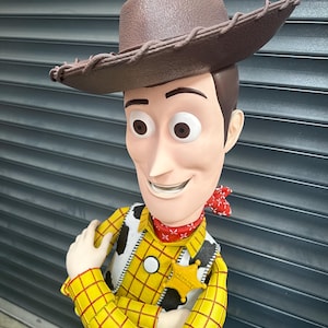 May include: A detailed figurine of Woody from Toy Story. The figure wears a brown cowboy hat, a yellow plaid shirt, a cow-print vest, a red bandana, and a sheriff's badge. The figure has a smiling expression.