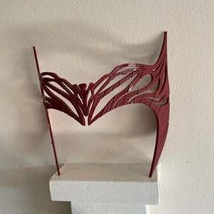 May include: A red plastic headband with a detailed, intricate design. The headband is displayed on a white shelf.