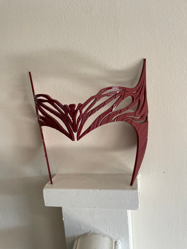 May include: A red plastic headband with a detailed, intricate design. The headband is displayed on a white shelf.