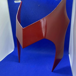 May include: A glossy, crimson-red headpiece with a distinctive angular design. The headpiece features a central, rectangular element and two pointed side pieces. The overall shape suggests a superhero or cosplay accessory.