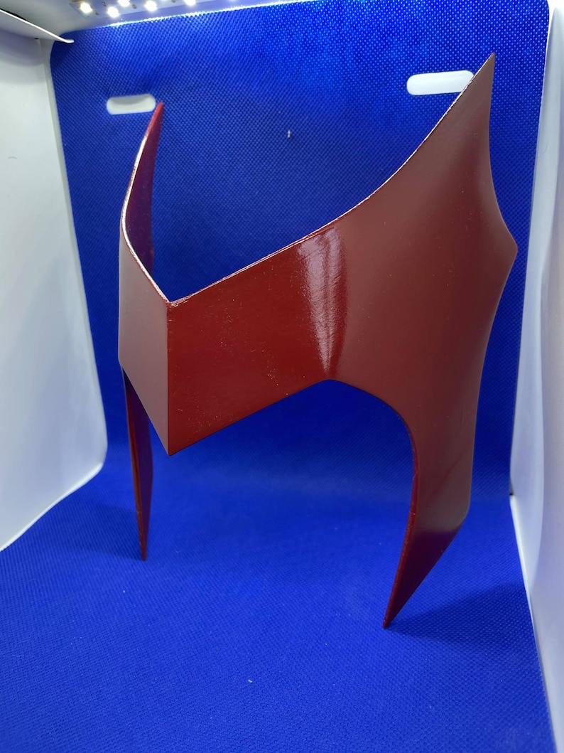 May include: A glossy, crimson-red headpiece with a distinctive angular design. The headpiece features a central, rectangular element and two pointed side pieces. The overall shape suggests a superhero or cosplay accessory.
