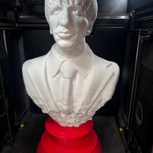 May include: A white bust of a man with a suit and tie, resting on a tiered red base. The bust features detailed facial features and hair. The sculpture is likely a 3D print.