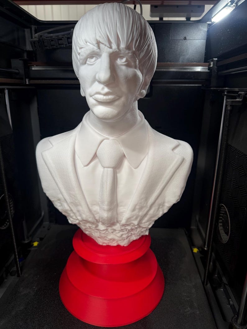 May include: A white bust of a man with a suit and tie, resting on a tiered red base. The bust features detailed facial features and hair. The sculpture is likely a 3D print.
