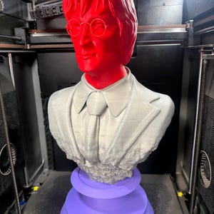May include: A 3D-printed bust of a man with red hair and glasses, wearing a gray suit and tie, atop a purple tiered base. The bust is in a 3D printer.