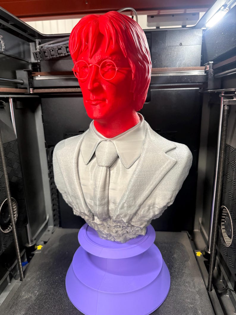 May include: A 3D-printed bust of a man with red hair and glasses, wearing a gray suit and tie, atop a purple tiered base. The bust is in a 3D printer.