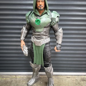 May include: A detailed action figure of a character in a green and silver armored suit with a hooded cowl. The figure has a stern expression and is holding a silver gauntlet. The armor features intricate detailing and a green circular emblem on the chest.