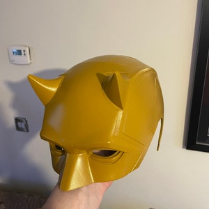 May include: A yellow plastic Daredevil mask with horns and eye holes. The mask is held in a hand.