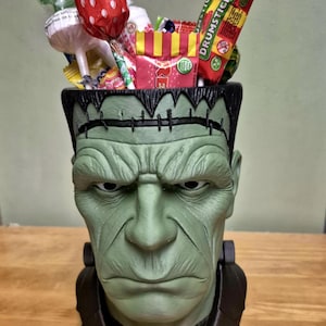 Puede incluir: Un recipiente verde con forma de cabeza de Frankenstein lleno de varios caramelos. El recipiente tiene una parte superior y un cuello negros. Los caramelos incluyen piruletas, dulces envueltos y un caramelo Drumstick. El recipiente est&aacute; sobre una superficie de madera.