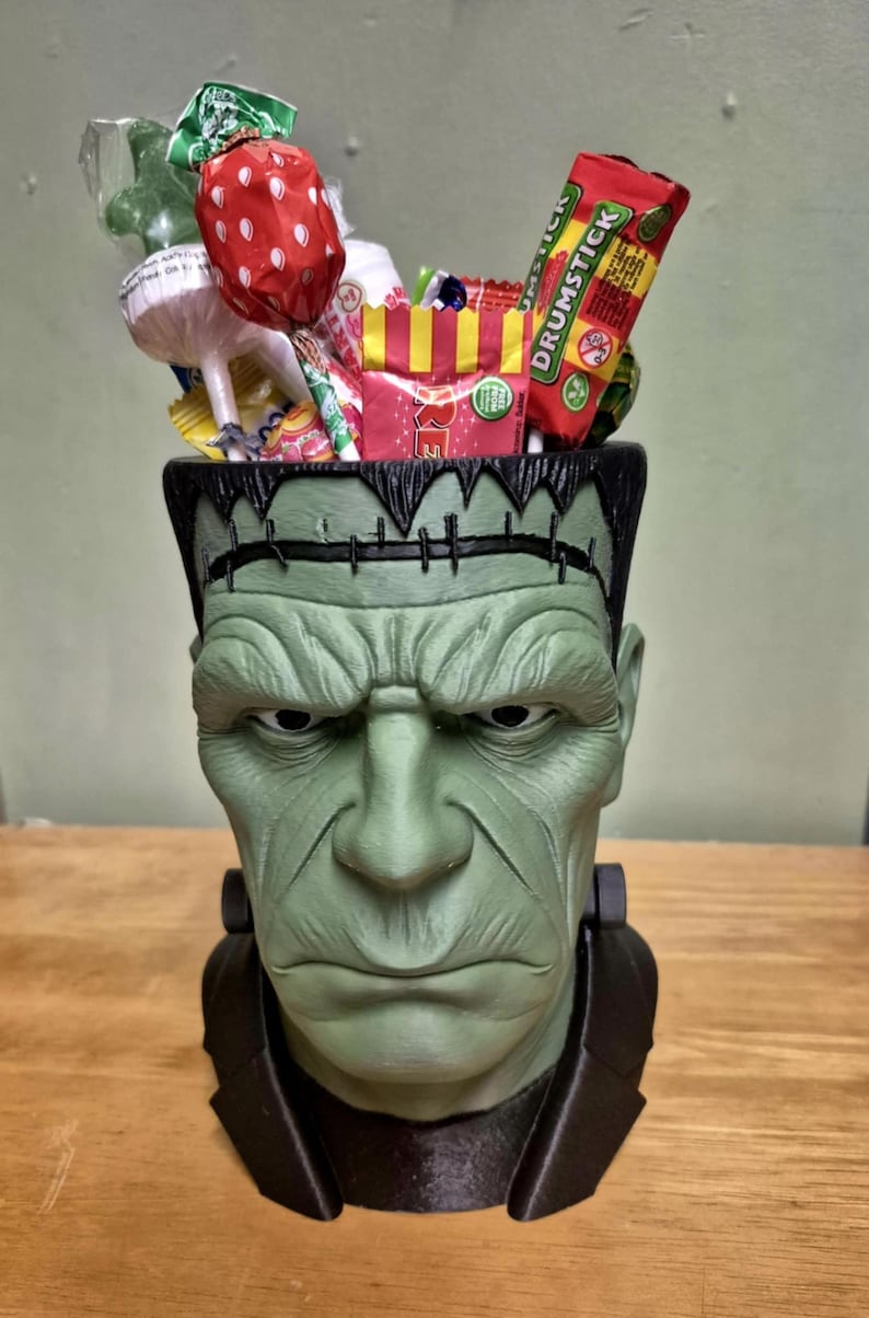 Puede incluir: Un recipiente verde con forma de cabeza de Frankenstein lleno de varios caramelos. El recipiente tiene una parte superior y un cuello negros. Los caramelos incluyen piruletas, dulces envueltos y un caramelo Drumstick. El recipiente est&aacute; sobre una superficie de madera.