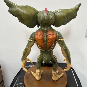 May include: A detailed sculpture of a Gremlin, viewed from the back. The figure is primarily green with orange and tan accents. It has large, textured ears, a spiky red crest, and sharp claws. The sculpture is mounted on a brown circular base.