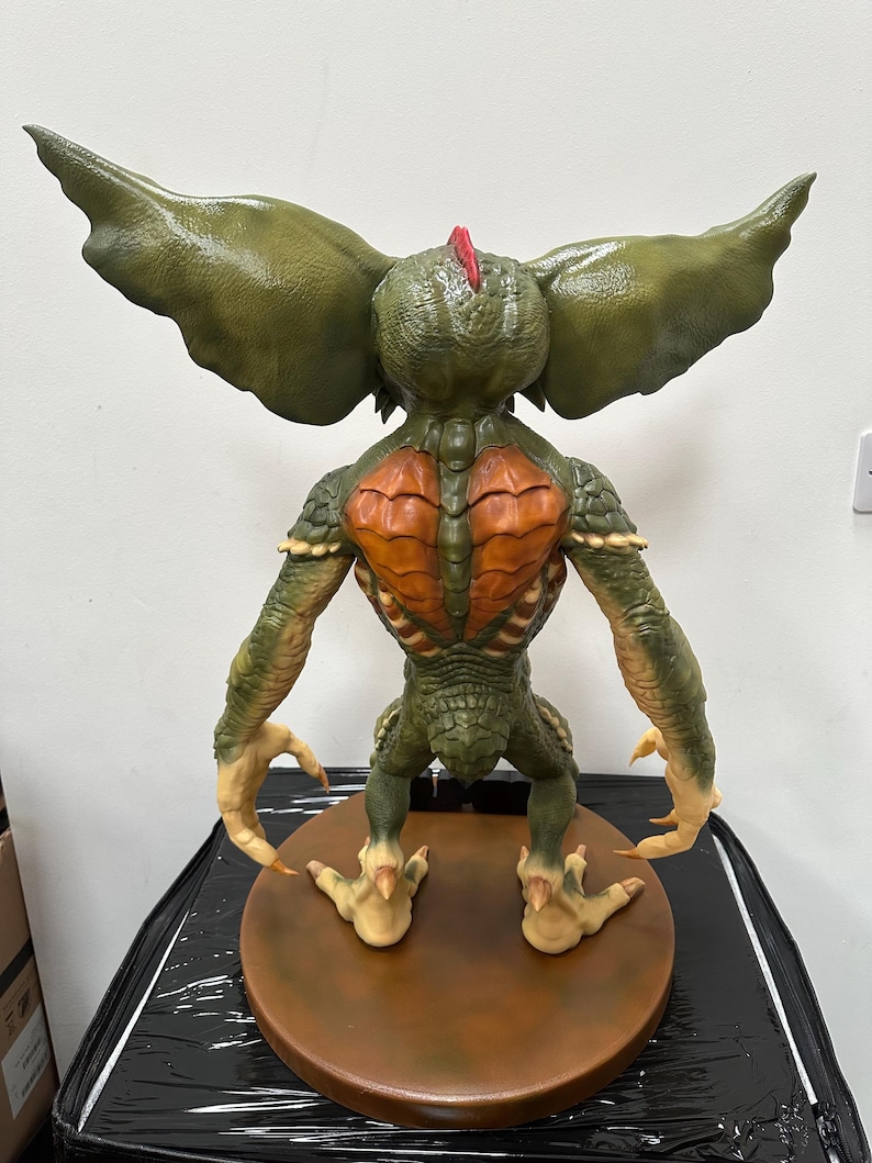 May include: A detailed sculpture of a Gremlin, viewed from the back. The figure is primarily green with orange and tan accents. It has large, textured ears, a spiky red crest, and sharp claws. The sculpture is mounted on a brown circular base.