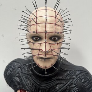 May include: A bust of a character with a pale complexion, adorned with nails embedded in the head. The face features dark eye makeup and a grid-like pattern. The character wears a black, textured garment with red accents.