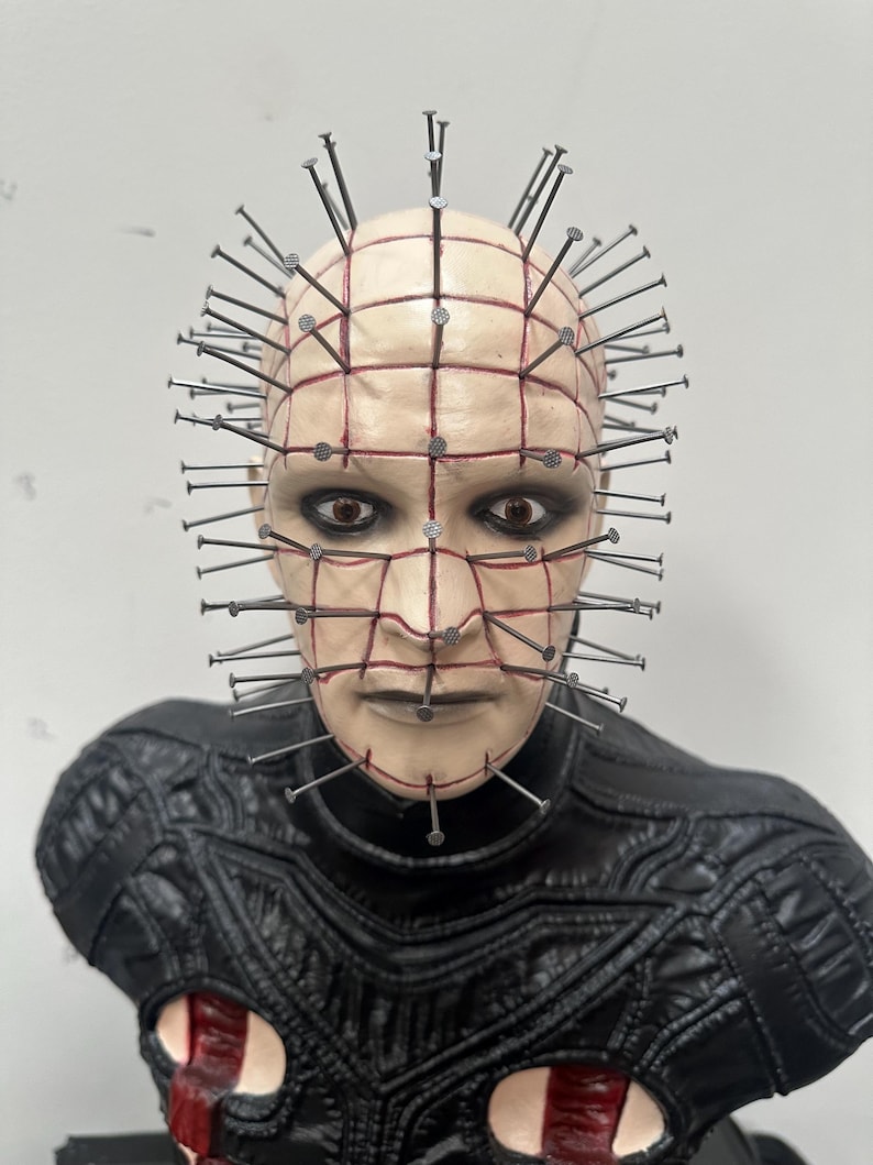 May include: A bust of a character with a pale complexion, adorned with nails embedded in the head. The face features dark eye makeup and a grid-like pattern. The character wears a black, textured garment with red accents.